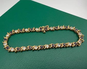 May include: A gold-tone tennis bracelet featuring an alternating pattern of round, dark blue sapphire-colored stones and clear, white diamond-colored stones set in a continuous S-link metal chain with a secure box clasp closure mechanism.