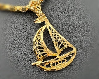 Peut inclure: Un collier doré avec un pendentif en forme de voilier. Le pendentif présente un design ajouré avec des détails complexes. Le collier est posé sur une surface sombre et texturée, mettant en valeur l'éclat et le savoir-faire du bijou.