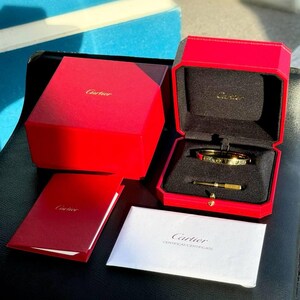 May include: A gold Cartier Love bracelet in its red box with a white certificate card. The box has a black interior and the bracelet is resting on a black cushion. The certificate card has the text 'Cartier CERTIFICAT-CERTIFICATE'.