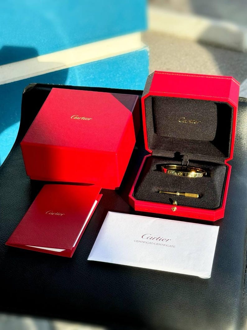 May include: A gold Cartier Love bracelet in its red box with a white certificate card. The box has a black interior and the bracelet is resting on a black cushion. The certificate card has the text 'Cartier CERTIFICAT-CERTIFICATE'.