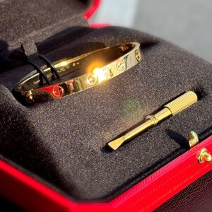 May include: A gold love bracelet with a screw design on a black velvet background inside a red box with a gold screwdriver.