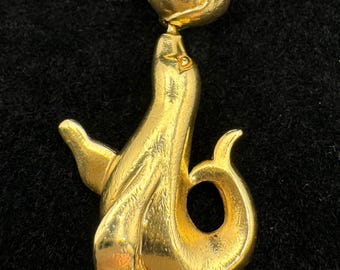 May include: A gold-toned pin featuring a stylized seal balancing a ball. The seal is depicted in a graceful pose, with its body curved and its flippers extended. The pin has a classic, elegant design, suitable for adding a touch of sophistication to any outfit.