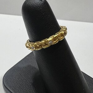 Puede incluir: Un anillo dorado con un patr&oacute;n repetitivo de eslabones ornamentados y peque&ntilde;as gemas transparentes. El anillo se muestra en un soporte negro. El dise&ntilde;o del anillo es detallado y parece ser vintage o antiguo.