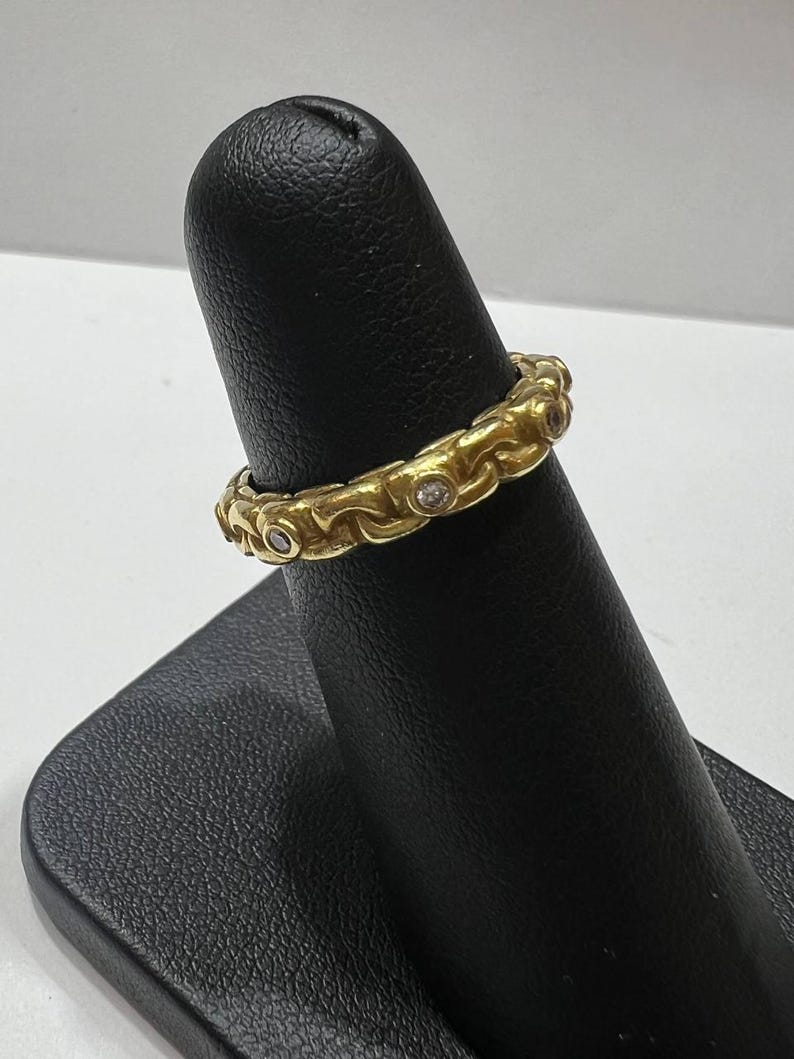 Puede incluir: Un anillo dorado con un patr&oacute;n repetitivo de eslabones ornamentados y peque&ntilde;as gemas transparentes. El anillo se muestra en un soporte negro. El dise&ntilde;o del anillo es detallado y parece ser vintage o antiguo.