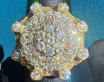 Peut inclure: Une bague dorée avec une large face circulaire, incrustée de nombreux petits diamants étincelants. Le design de la bague présente un groupe central de pierres entouré de cercles concentriques de gemmes, avec des accents hexagonaux sur le bord.