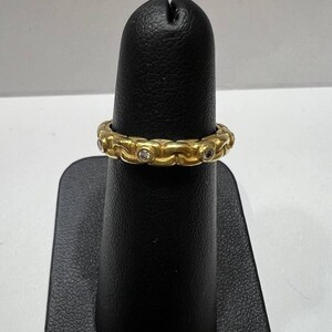 Puede incluir: Un anillo dorado con un dise&ntilde;o de eslabones de cadena repetidos. Peque&ntilde;as piedras preciosas transparentes est&aacute;n engastadas en los eslabones. El anillo se exhibe en un soporte negro. Es una joya.
