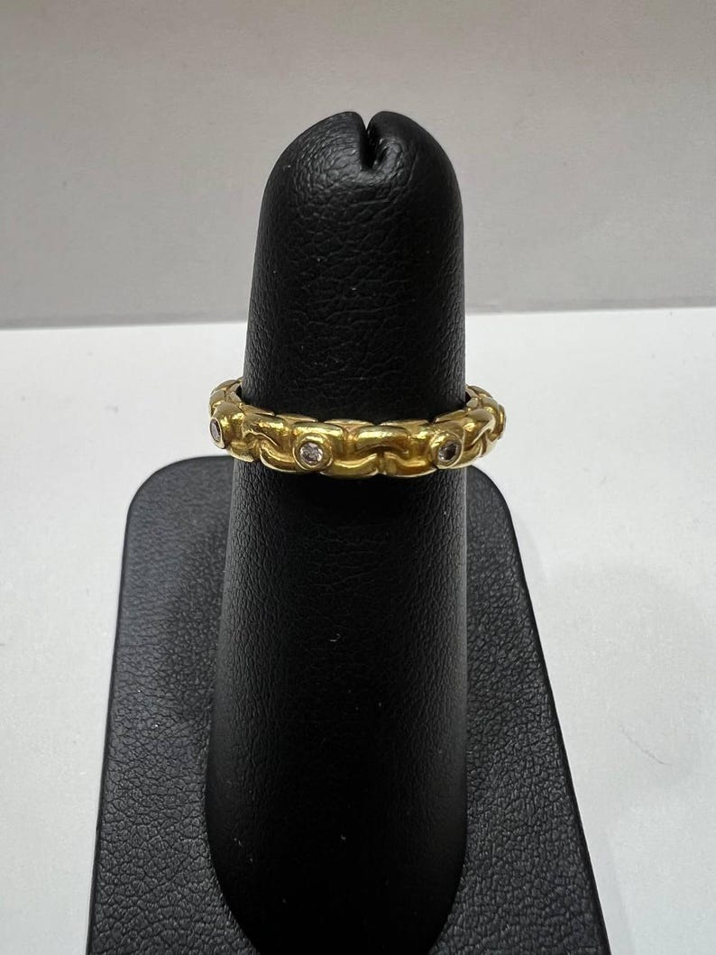 Puede incluir: Un anillo dorado con un dise&ntilde;o de eslabones de cadena repetidos. Peque&ntilde;as piedras preciosas transparentes est&aacute;n engastadas en los eslabones. El anillo se exhibe en un soporte negro. Es una joya.