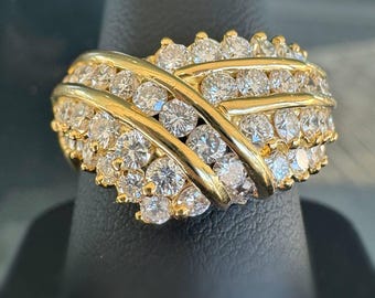 May include: A gold-toned ring with a cluster of clear, round gemstones. The ring features a crisscross design across the top, with multiple rows of gemstones. The ring is displayed on a black surface.