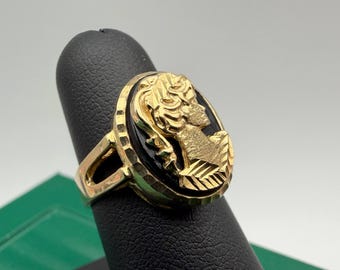 May include: A gold ring featuring an oval black stone inlay with a raised gold cameo profile of a person. The gold band has a textured, faceted edge surrounding the central stone, and the metal surface displays a polished, reflective finish.