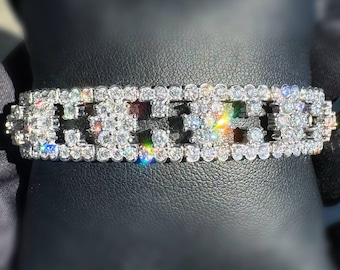 Peut inclure: Un bracelet argenté étincelant orné de nombreuses pierres claires et scintillantes. Le bracelet de forme rectangulaire est posé sur une surface noire. Les pierres reflètent la lumière, créant un affichage brillant.