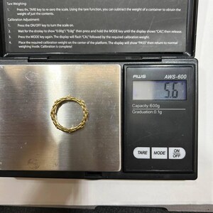Puede incluir: Un anillo dorado con un dise&ntilde;o tejido descansa sobre una balanza digital. La balanza muestra un peso de 5,6. La balanza tiene botones para TARA, MODO y ENCENDIDO/APAGADO. El texto en la balanza dice AWS-600, Capacidad: 600g, Graduaci&oacute;n: 0,1g.