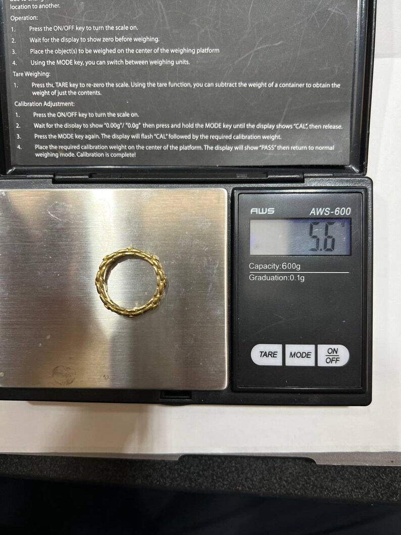 Puede incluir: Un anillo dorado con un dise&ntilde;o tejido descansa sobre una balanza digital. La balanza muestra un peso de 5,6. La balanza tiene botones para TARA, MODO y ENCENDIDO/APAGADO. El texto en la balanza dice AWS-600, Capacidad: 600g, Graduaci&oacute;n: 0,1g.