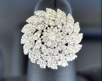 May include: A dazzling, silver-toned ring featuring a floral design. The ring is encrusted with numerous small, sparkling gemstones, arranged in a layered, petal-like pattern. The ring is set against a dark background, highlighting its brilliance.