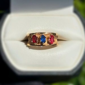 May include: A gold ring featuring three oval gemstones: two red and one blue. The ring has a textured band and is presented in a white jewellery box.