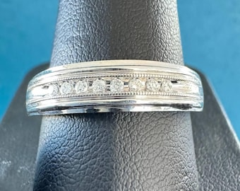 Peut inclure: Une bague en argent ornée d'une rangée de petits pierres précieuses transparentes. La bague présente un motif détaillé et texturé sur les bords. La bague est présentée sur un support noir sur fond bleu. Bijoux.