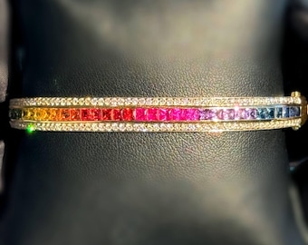 Peut inclure: Un bracelet en or orné de pierres précieuses taillées carrées aux couleurs de l'arc-en-ciel. Le bracelet présente une rangée de diamants au-dessus et en dessous des pierres colorées. Les pierres précieuses passent du jaune au rouge, rose, violet et bleu.
