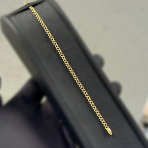 May include: A delicate gold chain bracelet with a curb link design. The bracelet is displayed on a black surface. The gold color of the bracelet contrasts with the dark background, highlighting its intricate details. The bracelet is approximately 7 inches long.