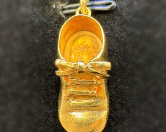 May include: A gold-colored baby shoe pendant with a bow detail. The pendant has a loop at the top for a chain. The shoe has laces and a textured surface. The pendant is likely made of gold or gold-plated metal.