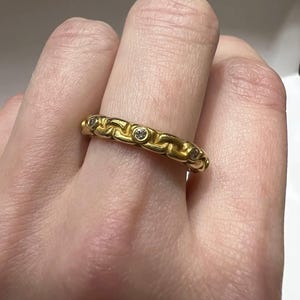 Puede incluir: Un anillo dorado con un dise&ntilde;o de eslabones de cadena. El anillo presenta peque&ntilde;as piedras preciosas transparentes engastadas en los eslabones. El anillo se lleva en un dedo, mostrando sus intrincados detalles y artesan&iacute;a. Es una joya.