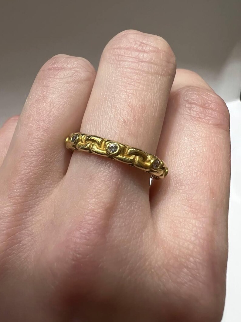 Puede incluir: Un anillo dorado con un dise&ntilde;o de eslabones de cadena. El anillo presenta peque&ntilde;as piedras preciosas transparentes engastadas en los eslabones. El anillo se lleva en un dedo, mostrando sus intrincados detalles y artesan&iacute;a. Es una joya.