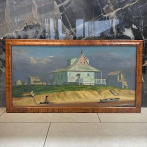 Puede incluir: Pintura al óleo enmarcada que representa una escena costera con una casa grande en una pequeña colina. Una persona está sentada en la playa cerca de un bote. El cielo es una mezcla de azul y gris, lo que sugiere un día nublado. Ambiente vintage.