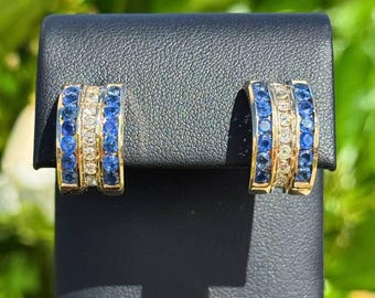 May include: A pair of gold-toned earrings with a curved design. Each earring features three rows of gemstones: blue, clear, and blue. The earrings are displayed on a black surface against a blurred green background.