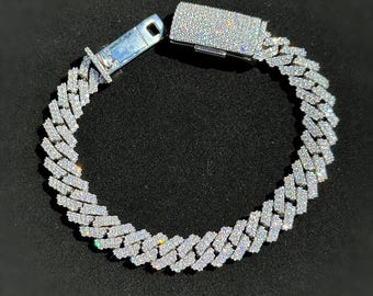 May include: A dazzling, silver-toned bracelet with a cuban link design. The bracelet is encrusted with numerous small, sparkling stones, creating a brilliant, eye-catching effect. The clasp is also covered in the same shimmering stones. The jewelry is displayed against a dark background.
