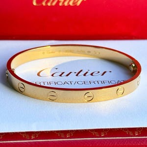 May include: A gold Cartier Love bracelet with a screw design on a white surface. The bracelet is in a red Cartier box with the Cartier logo on the top.