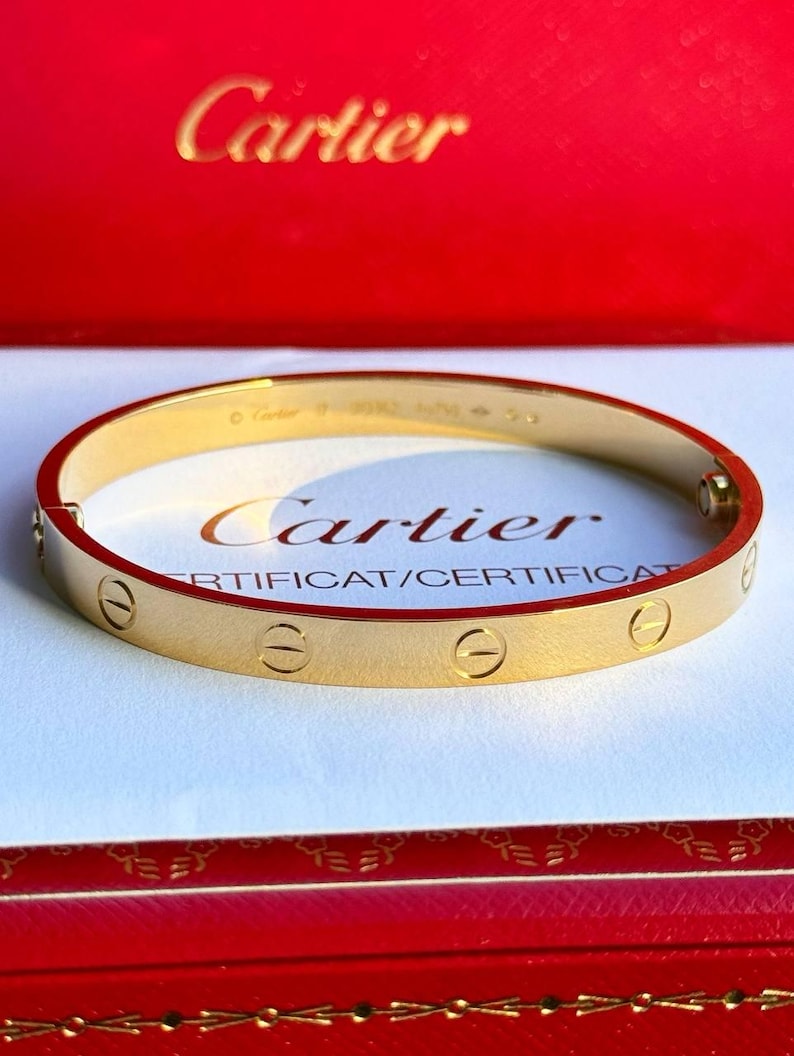May include: A gold Cartier Love bracelet with a screw design on a white surface. The bracelet is in a red Cartier box with the Cartier logo on the top.