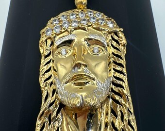 May include: A gold-toned pendant featuring a detailed face of Jesus with a crown of thorns. The crown and eyes are accented with clear, sparkling stones. The pendant includes a decorative bail at the top, also set with three clear stones.