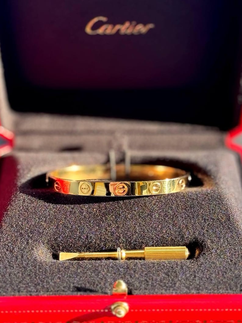 May include: A gold Cartier Love bracelet in its red box with a gold screwdriver. The box has the Cartier logo on the top.