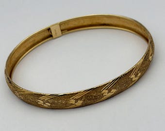 May include: A gold-toned hinged bangle bracelet featuring a textured surface with repeating geometric patterns of diagonal lines and small squares. The bracelet has a polished interior and a secure metal clasp mechanism visible on the side.
