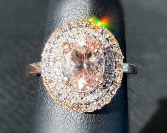 Peut inclure: Une bague en or rose ornée d'une grosse pierre ovale rose pâle, entourée de deux halos de petites pierres claires et couleur champagne. La bague est posée sur un fond noir, avec la lumière qui se reflète sur les pierres.