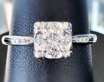 Peut inclure: Une bague en argent avec un sertissage carré et orné. Le centre présente une grosse pierre précieuse ronde et transparente, entourée de petites pierres. La bande comporte des petites pierres supplémentaires de chaque côté, créant un effet scintillant. La bague est présentée sur un fond sombre.