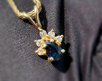 Peut inclure: Un collier en or avec un pendentif orné d'une pierre précieuse ovale bleu foncé. La pierre est entourée de petites pierres précieuses claires. La chaîne du collier est fine et tressée.