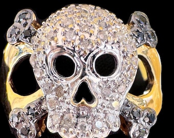 Peut inclure: Une bague en forme de crâne avec une bande dorée et un motif de crâne incrusté de petits diamants étincelants. Le crâne a des orbites noires et un nez noir. Deux tibias croisés sont derrière le crâne, avec des gemmes noires aux extrémités.