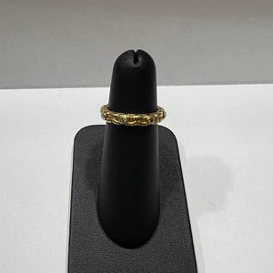 Puede incluir: Un anillo dorado con un patr&oacute;n repetitivo de peque&ntilde;as piedras redondas y transparentes. El anillo se exhibe en un soporte negro sobre un fondo blanco. El anillo parece estar hecho de metal.