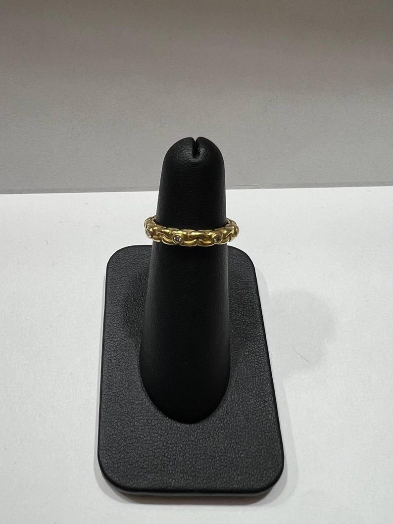 Puede incluir: Un anillo dorado con un patr&oacute;n repetitivo de peque&ntilde;as piedras redondas y transparentes. El anillo se exhibe en un soporte negro sobre un fondo blanco. El anillo parece estar hecho de metal.