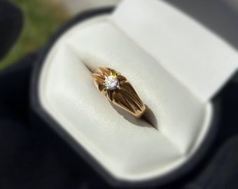 Peut inclure: Une bague en or avec un diamant central, sertie dans un motif stylisé en forme d'éclat de soleil. La bague est présentée dans un écrin à bijoux blanc et rembourré, avec un extérieur noir. La bande de la bague présente un motif géométrique texturé.