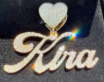 May include: A gold pendant with the name "Kira" in a cursive font, encrusted with small, sparkling stones. Above the name is a heart-shaped charm, also covered in the same stones. The pendant is set against a dark background.