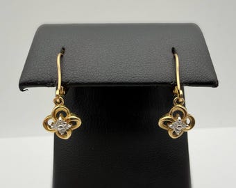 May include: A pair of gold drop earrings featuring a four-leaf clover design. Each earring has a central cluster of small, clear, sparkling stones set within the gold clover shape, suspended from a curved gold wire hook for secure attachment.