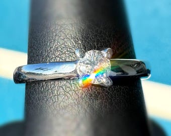 Peut inclure: Une bague en argent avec un diamant clair taillé en rond. Le diamant est le point central, reflétant un arc-en-ciel de couleurs. La bague est posée sur une surface noire avec un fond bleu.