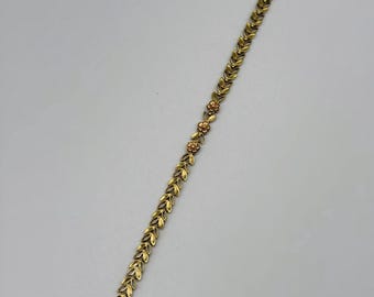 May include: A gold-toned metal bracelet featuring a repeating leaf-shaped link pattern. The design includes a small section of three integrated flower-shaped links near the center. The bracelet is finished with a secure clasp at one end.