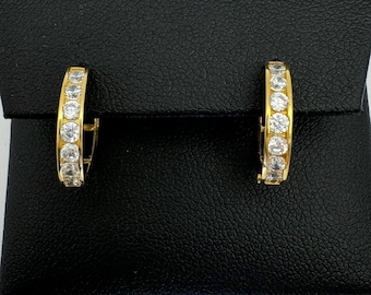 May include: A pair of gold-toned hoop earrings, each encrusted with a row of clear, sparkling stones. The earrings are displayed on a black, textured surface, creating a contrast that highlights their brilliance. The design is classic and elegant.