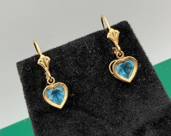 May include: A pair of gold drop earrings featuring heart-shaped blue gemstones. Each earring has a gold hook wire and a small gold setting that holds the faceted blue heart stone, creating a dangling design that catches the light.