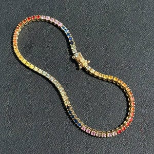 Puede incluir: Una pulsera dorada con una gama de piedras preciosas. Los colores van del rojo al naranja, amarillo, verde, azul y morado. La pulsera se asegura con un cierre.