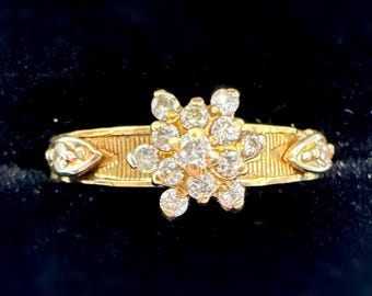 May include: A gold ring featuring a cluster of round-cut diamonds in a floral arrangement. The band has a textured design with decorative elements on each side of the diamond cluster. The ring is set against a dark background.