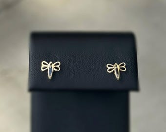 Peut inclure: Une paire de boucles d'oreilles clous en forme de libellule dorée. Chaque boucle d'oreille présente un motif de libellule délicat avec un petit accent bleu foncé. Les boucles d'oreilles sont présentées sur une surface noire.