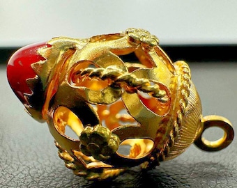 May include: A gold-colored pendant with a red gemstone. The pendant has an intricate openwork design with floral and rope-like details. It features a loop for hanging and is likely a piece of jewelry.