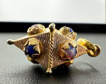May include: A gold-colored pendant with a starburst design. The pendant features blue and purple gemstones set within ornate gold settings. The piece has a textured surface and a small loop for a chain.
